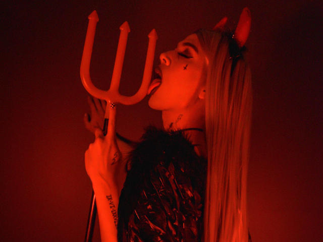 AlessandraFerrer with red lights, tongue out, holding a trident, wearing devil horns and a shiny black top. Tattoo on her wrist. Long blonde hair. Red and black.
