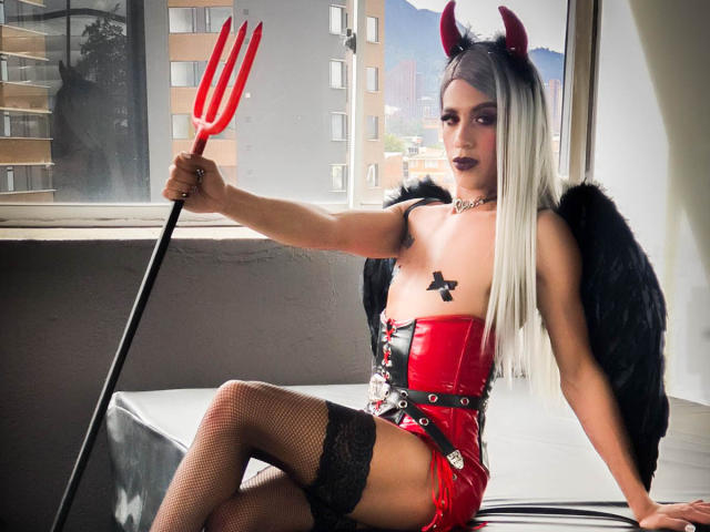 Alessandra Ferrer com chifres de demônio, asas pretas, corpete de látex vermelho, bojos de estrela pretos, meias de rede, segurando um tridente vermelho, sentada na cama, cabelo de branco para preto, batom escuro..