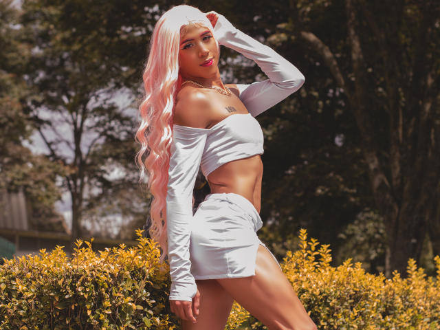 Alessandra Ferrer, con cabello rosa, un top corto blanco con un hombro descubierto y una minifalda a juego, mostrando sus glúteos tonificados y sus pequeños senos, de pie en un jardín iluminado por el sol..