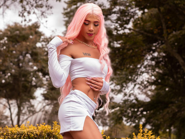 Alessandra Ferrer, avec une perruque rose, un crop top blanc à épaules dénudées et une jupe blanche, montrant ses seins, avec une main sur ses fesses, dans une forêt, portant un collier en or..