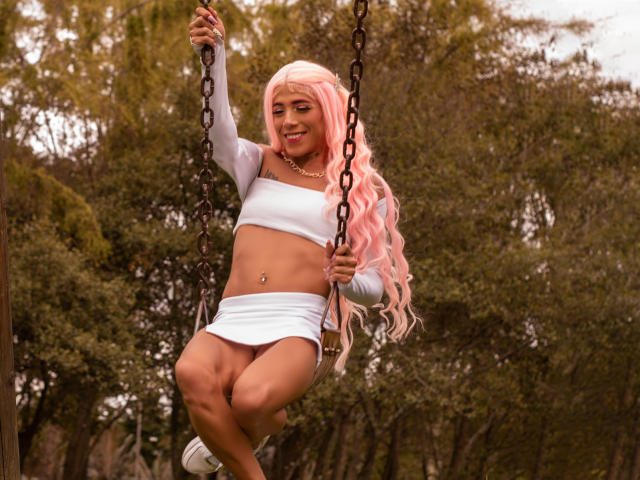 Alessandra Ferrer, com peruca rosa, cropped branco e minissaia, sentada em um balanço, segurando correntes, mostrando um piercing no umbigo, sorrindo e exibindo seus tênis brancos..