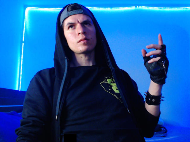 DreamHunterdz with a pensive look, wearing a black hoodie and backwards cap, pointing with a finger while wearing black fingerless gloves, in a blue-lit room.