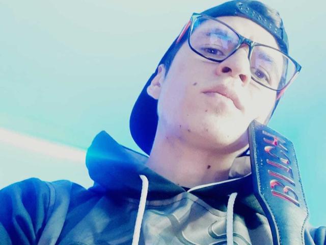 DreamHunterdz mit Brille, der eine schwarze Mütze und einen Kapuzenpullover trägt, mit ein paar Sommersprossen im Gesicht, der leicht grinsend nach unten schaut, blauer Himmel im Hintergrund..