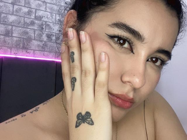 Le visage de Liana Bernocchi, avec ses cheveux foncés, ses yeux marron et son eye-liner ailé. Elle se couvre la bouche avec sa main tatouée, qui révèle des tatouages de papillons et une petite croix. Elle est nue..