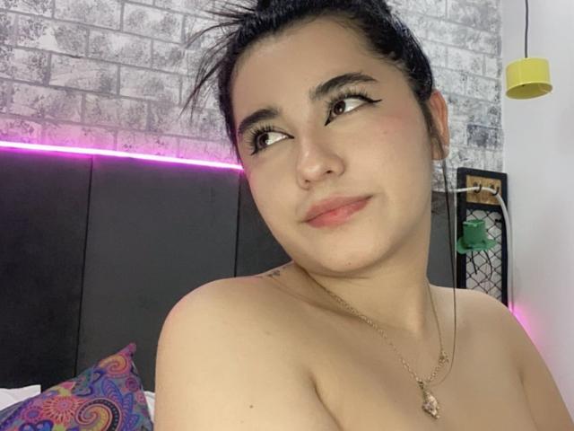 Liana Bernocchi tem cabelo escuro preso em um coque desarrumado, delineador e um leve sorriso, com os ombros descobertos e um colar, olhando para o lado. Seus seios não estão visíveis..