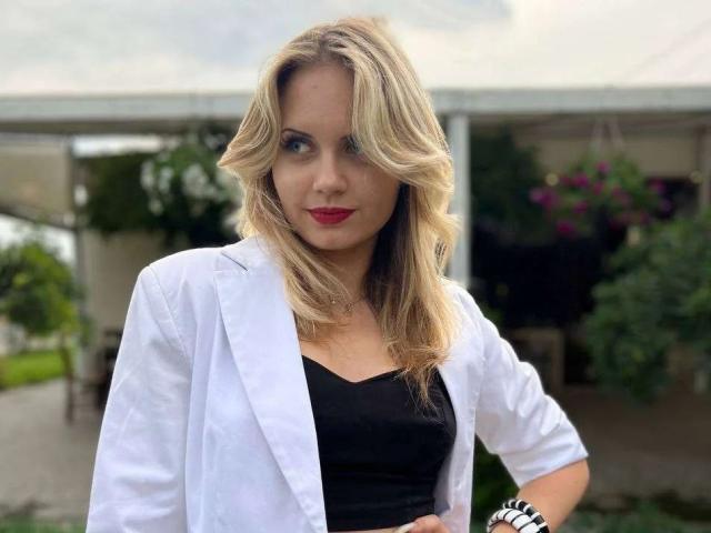 LoriVal, rubia, con cabello ondulado, lápiz labial rojo, bata de laboratorio blanca, top negro y pulsera en la muñeca, de pie con una mano en la cadera, mirando a la cámara. Los senos están cubiertos por el top negro..