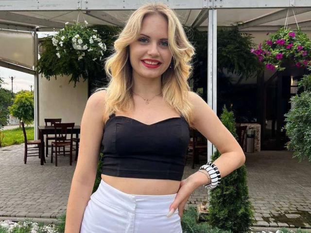 LoriVal, con cabello rubio ondulado, lápiz labial rojo, top negro sin tirantes, pantalones blancos y pulsera negra y blanca, sonriendo, con la mano en la cadera, debajo de una carpa blanca con flores colgantes..
