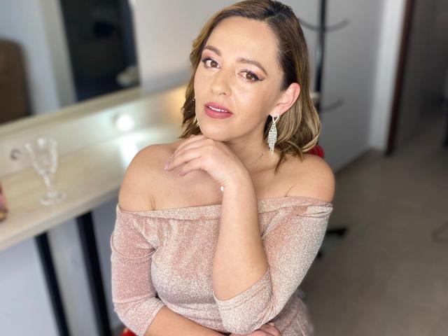 AmandaHotL with shoulderless pink sparkly top, brown wavy hair, pink lipstick, looking at camera, hand on chin, earrings, subtle makeup, white background, kitchen counter on left.