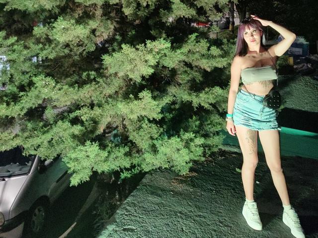 Samantha Morgan con un top verde de tubo, pantalones cortos de cintura alta a juego, zapatillas verdes, cabello morado, de pie debajo de un árbol grande, con una mano en la cabeza, tatuaje visible en el muslo, un coche parcialmente visible a la izquierda..