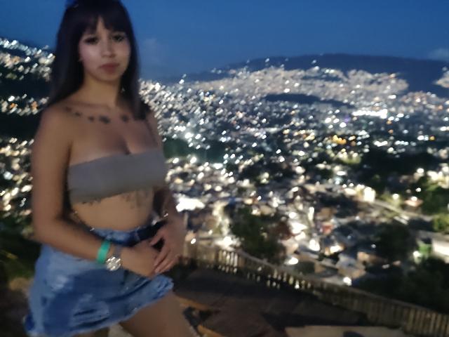 Samantha Morgan, con senos de tamaño medio, vestida con un top sin tirantes y una falda vaquera, de pie al aire libre por la noche, con luces de la ciudad en el fondo, cabello oscuro, collar y una pulsera verde. Foto borrosa..