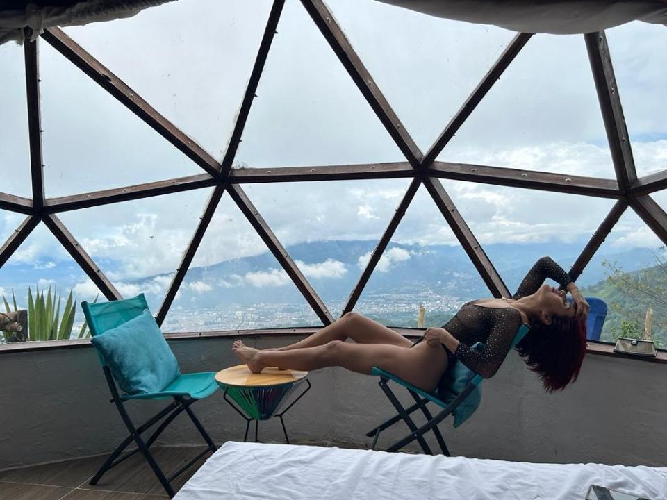 ErzaScarlet est assise sur une chaise turquoise, vêtue d'un haut noir transparent à manches longues, sans pantalon, les jambes relevées, dévoilant ses fesses, avec ses cheveux roux, dans une pièce en verre aux motifs géométriques..