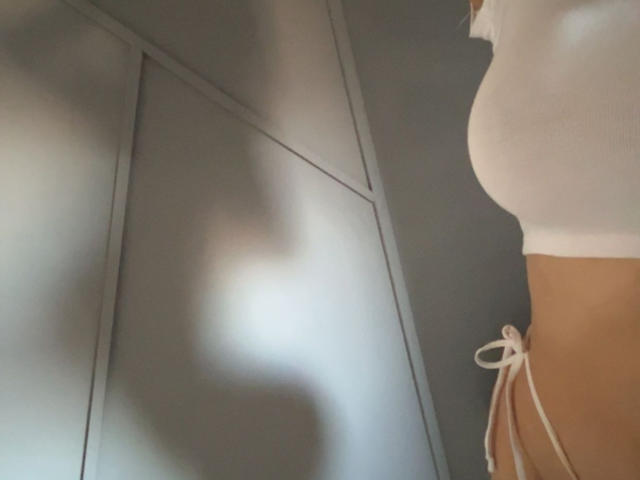Le profil de Christine Lagarde, vêtue d'un haut blanc, montrant ses petites seins et un bas de maillot de bain blanc en string, sur un fond gris et ombragé..