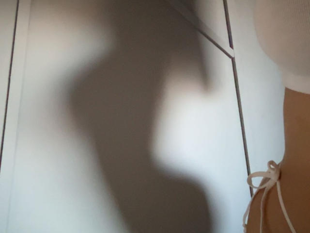 L'ombre des fesses et des seins de Christine Lagarde projetée sur un mur blanc, avec un ruban blanc sur le côté droit et un objet circulaire blanc dans le coin supérieur droit..