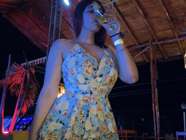 NicoColeman with big boobs, wearing a floral dress, drinking from a glass, under a wooden roof, blue light, red palm leaves, wristbands, night, holding her glass up.