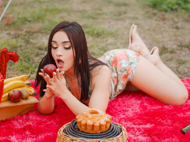 AngelCutie con lunghi capelli scuri, in un costume intero a fiori, su una coperta rossa e pelosa, mentre mangia una mela, sdraiata a pancia in giù, con una ciambella davanti, banane e mele su un vassoio, con la bocca aperta, che appare sexy..