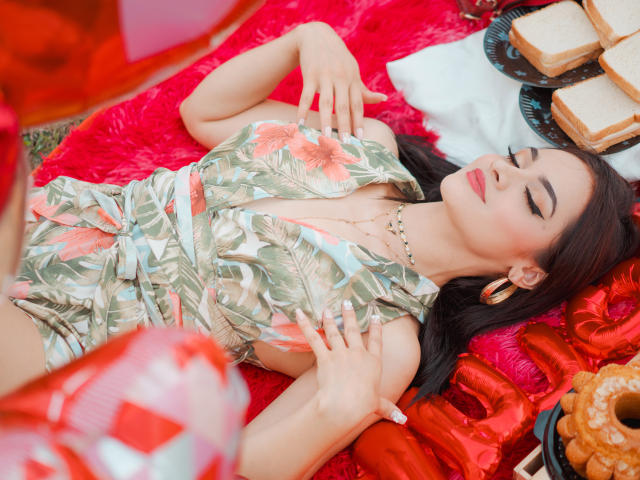 AngelCutie avec des cheveux foncés, les yeux fermés, portant une robe fleurie, allongée sur un tissu rouge, les mains sur la poitrine, avec des boucles d'oreilles dorées et un collier, avec des sandwichs et des beignets à proximité..