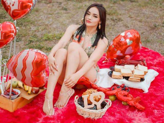 AngelCutie met borsten in een groene kanten bh, zittend op een rode, pluizige deken, benen gekruist, omringd door hartvormige ballonnen, koekjes, fruit en een mandje met pure chocolade..