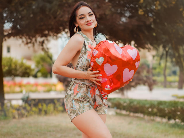 AngelCutie mit dunklem Haar, trägt einen grünen, mit Blumen bedruckten Overall, hält einen roten, herzförmigen Luftballon mit rosa Herzen, steht in einem Park, lächelt und zeigt ihre Brüste..