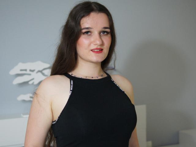 KateUnique mit langen, braunen Haaren, rotem Lippenstift, schwarzem Top ohne Ärmel, das ihre Brüste zeigt, heller Haut, leichtem Lächeln, Tattoo am Hals, weiße Wand, weiße Möbel im Hintergrund..