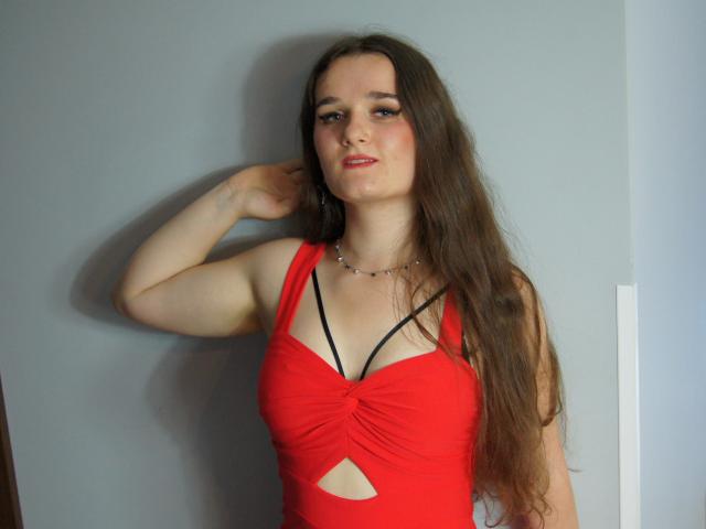 KateUnique with long brown hair, red dress, black strappy bra, small necklace, standing against gray wall, hand raised, looking at camera, red lips, light skin, slight smile.
