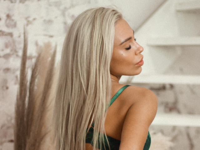 KateLilly, con lunghi capelli biondi, reggiseno verde, labbra carnose e pelle luminosa, guarda oltre la spalla, mostrando la schiena e i suoi piccoli seni sodi. Illuminazione soffusa e calda..