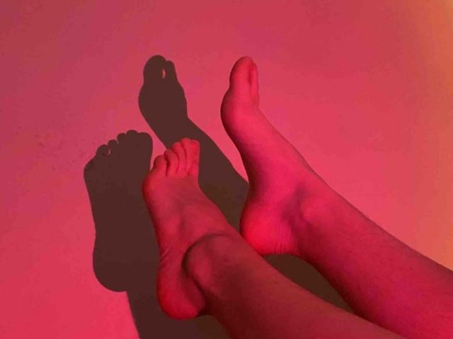 SarahyJhones' legs with feet crossed, showing shadows on a pink-tinged wall. Her toes are slightly curled, and her soles are visible. The shadow of her feet is dark.