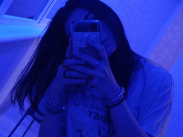 Zanoza, con cabello largo y oscuro, rostro iluminado en azul, sosteniendo un teléfono frente a ella, vestida con una camisa y brazaletes, tomando una selfie, con un anillo visible en la mano izquierda, mirando hacia abajo, ligeramente borroso..