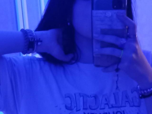 Zanoza's hand grijpt haar borsten door een wit shirt, en ze maakt een spiegelzelfportret met blauw licht. Haar andere hand houdt de telefoon vast, waarbij haar armbanden zichtbaar zijn..