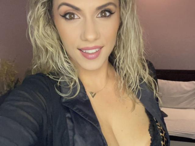 LucilleXSugar, com cabelo loiro ondulado, vestindo uma camisa preta, revelando o colo com um sutiã de estampa de leopardo, sorrindo com batom rosa, olhos castanhos e um pequeno colar em formato de triângulo..