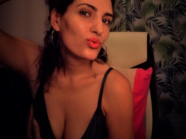 AddictedToUTS with pouty lips, wearing a black lace bra, showing her tits, and large hoop earrings, has dark hair and brown skin, in a dimly lit room.