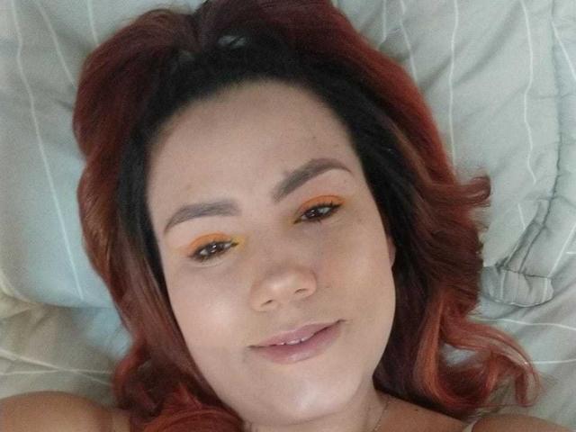 Eine Sharon Grey mit roten Haaren, gelbem Lidschatten und einem leichten Lächeln, liegt auf einem Bett mit weißen Kissen. Ihr Gesicht ist zentriert, und man sieht ihren entspannten Ausdruck..