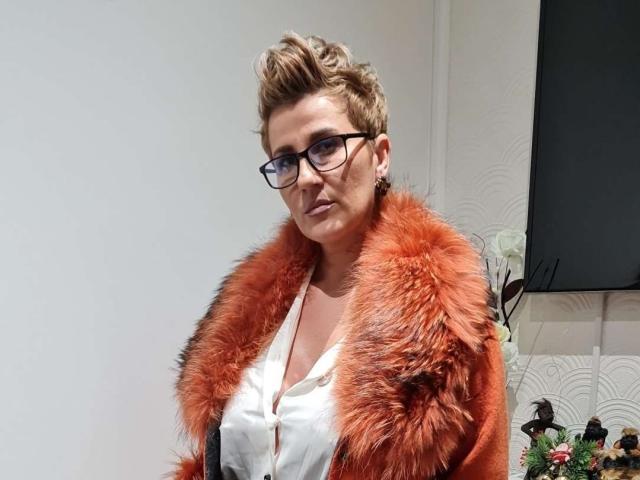 InanAIshtars with short, messy blonde hair, glasses, and a fluffy orange fur coat over a white shirt, looking slightly annoyed. Boobs are partially visible under the coat.