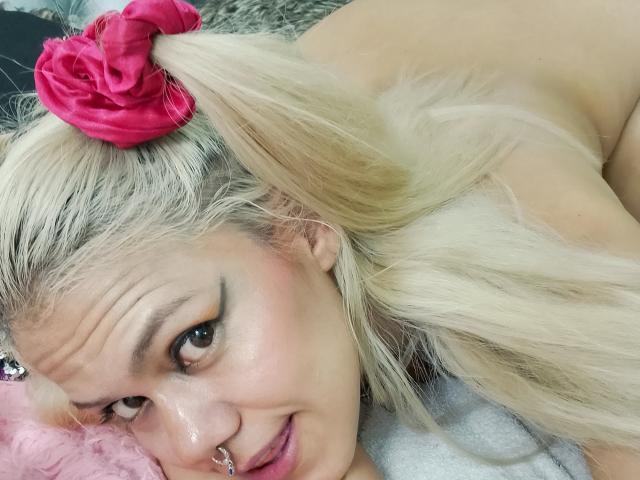 TarahDustin with blonde pigtails, pink scrunchie, nose ring, and black eyeliner, lying down, smiling, showing a bit of her shiny ass, on a pink pillow.