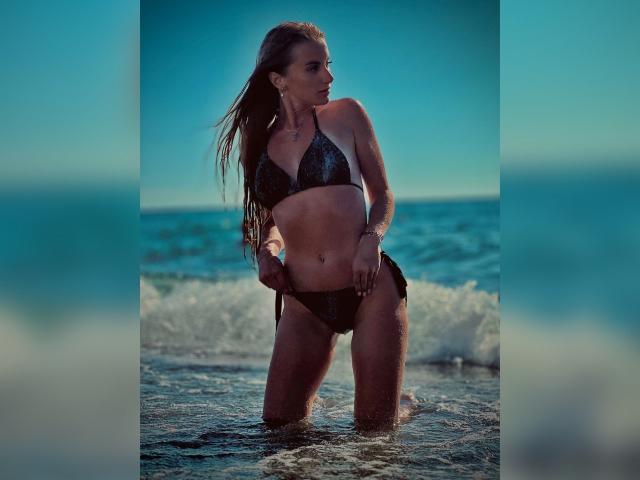 BlondyOneSex kniet im flachen Wasser, trägt einen schwarzen, glitzernden Bikini, hat nasses Haar, definierte Bauchmuskeln und kleine Brüste, vor einem blauen Ozean im Hintergrund..