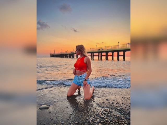 BlondyOneSex kniet im feuchten Sand, trägt ein rotes Crop-Top und blaue Shorts, wobei ihr Hintern und ihre Brüste sichtbar sind, am Fuße eines hölzernen Piers bei Sonnenuntergang..