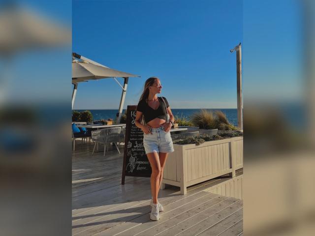 BlondyOneSex mit langen, braunen Haaren, schwarzem Crop-Top, hoch taillierten Jeansshorts, weißen Sneakers, stehend auf einem Holzboden, blauer Himmel, Ozean im Hintergrund, lächelnd, Hände an den Hüften..