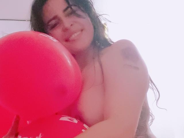 LindaSmith with long dark hair, holding a red balloon, smiling, topless, showing her big tits, and a small tattoo on her right shoulder, in a bright room.