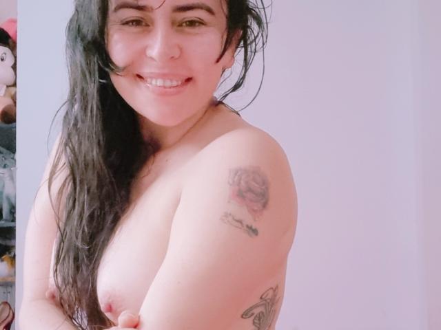 LindaSmith, topless, with long black hair, smiling, showing tattoos on her right arm and upper chest, medium-sized tits, light skin, and a cute nose ring.