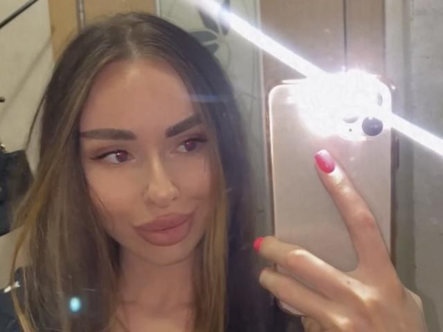 BellaMegan, con lunghi capelli castani, labbra piene rosate e smalto rosso, sta scattando un selfie allo specchio, tenendo in mano un iPhone rosa, con riflessi luminosi nella parte superiore destra..