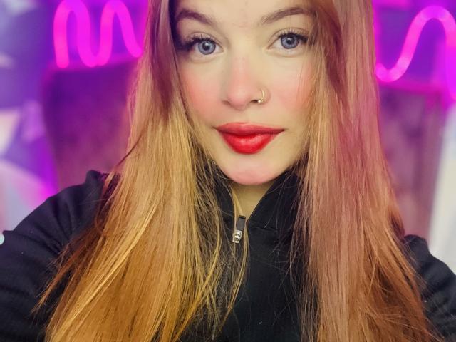Shantal, con cabello largo de color castaño rojizo, un piercing en la nariz y lápiz labial rojo, vestida con una chaqueta negra, mirando directamente a la cámara con ojos azules y una ligera sonrisa..