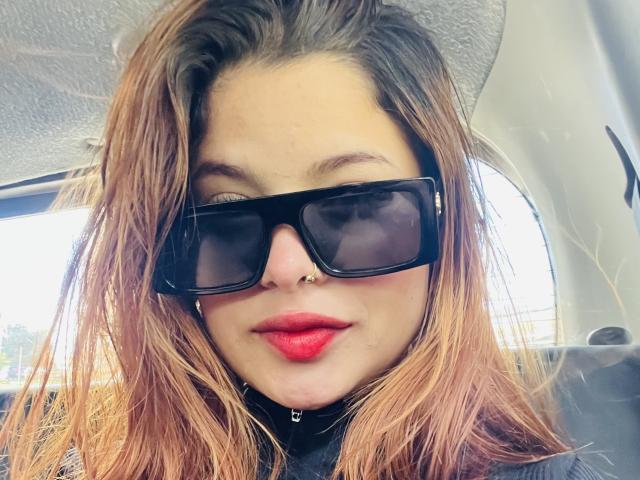 ShantalSummer has dirty blonde hair, black sunglasses, red lipstick, and a nose ring. She's wearing a black shirt with a zipper, sitting in a car.