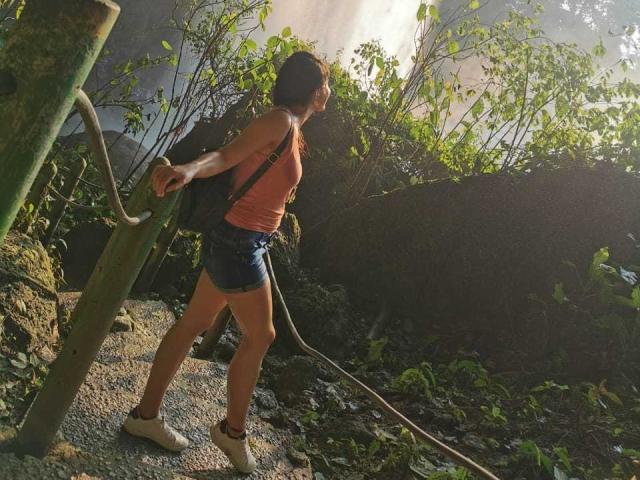 CheeryMia met bruine huid, een rood tanktopje, denim shorts, witte sneakers, een rugzak, die een touw vasthoudt, met zonlicht dat door de bladeren schijnt, een groene struik, een rots en een metalen paal in het zonlicht..