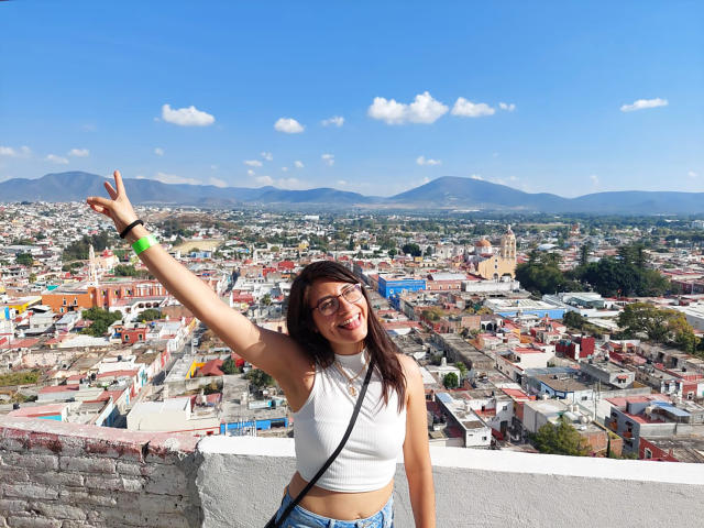 CheeryMia, avec des cheveux bruns, des lunettes, un débardeur blanc, un jean bleu, levant la main droite, souriant, avec des montagnes et une ville en arrière-plan, portant un bracelet vert, debout sur un mur de briques blanches..