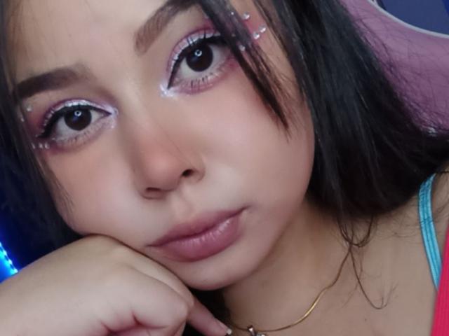 AlizeeTorres with pink eyeshadow, black eyeliner, and glitter, looking at camera with hand under chin, has pink hair clip, and a gold necklace, has small tits and a cute face.