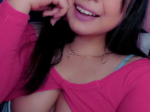 AlizeeTorres with a smile, wearing a pink top that reveals her large, round tits, her hand touching her cheek, long black hair, and a cross necklace.