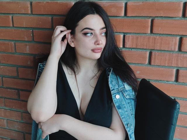 MilanaPrice, com pele clara, cabelo escuro e olhos azuis, vestindo uma blusa preta com decote em V e uma jaqueta jeans, está sentada encostada em uma parede de tijolos, com a mão no cabelo, olhando para o lado..