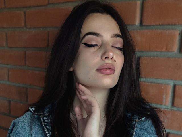 MilanaPrice tem cabelos longos e escuros, pálpebras rosadas e um piercing no nariz, com os olhos fechados e os lábios levemente curvados, vestindo uma jaqueta jeans e tocando o pescoço..