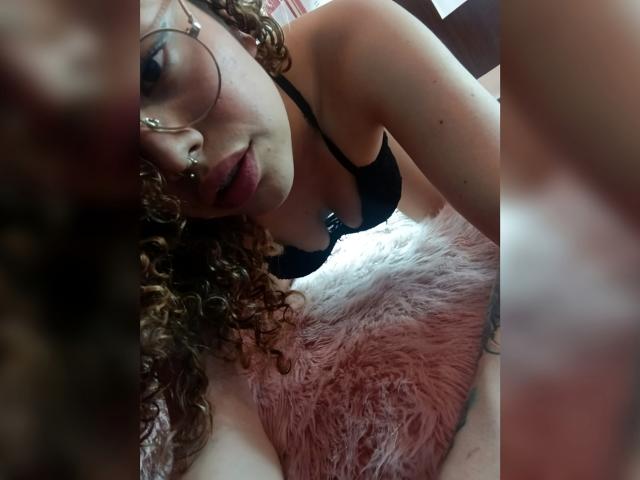 KaterinXChris mit lockigem, braunem Haar, Brille, Nasenring und schwarzem BH, lehnt sich an ein pinkes, flauschiges Kissen und zeigt ihre Brüste. Ihr Gesicht ist nah, sie schaut in die Kamera..