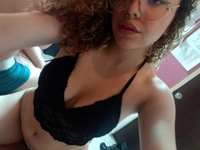KaterinXChris mit lockigem, braunem Haar, Brille, Nasenring, schwarzem Spitzen-BH, die ihre Brüste zeigt, und pinkem Lippenstift, in einem Selfie, mit ihrem Hintern in türkisfarbenen Unterwäsche..
