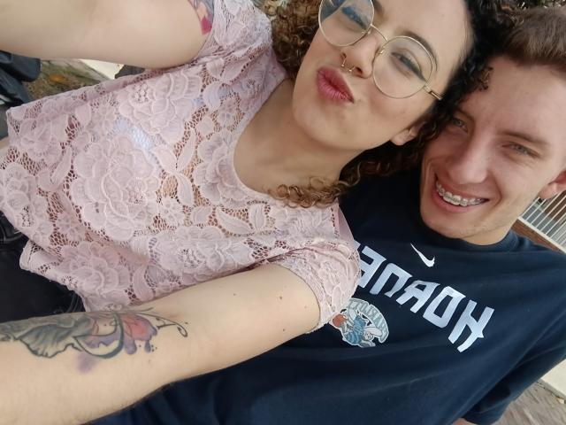 KaterinXChris met krullend bruin haar, een bril, een roze kanten top en een tatoeage op de arm. KaterinXChris draagt een zwarte Nike shirt, glimlacht en maakt een selfie. De borsten en de tatoeage op de arm van KaterinXChris zijn zichtbaar..